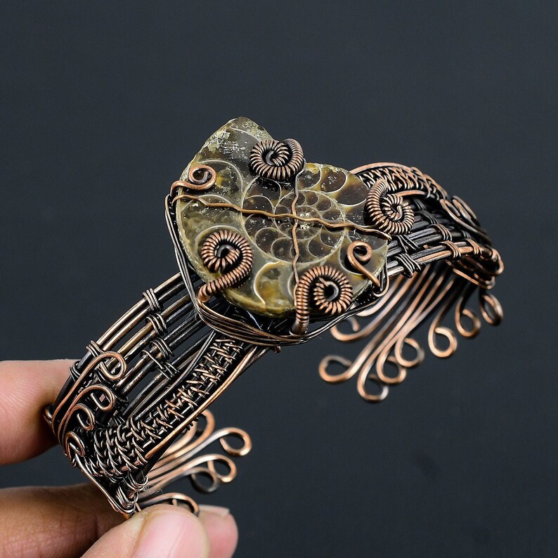 May include: A copper wire wrapped bracelet featuring a brown and gold ammonite fossil as the centrepiece. The bracelet is woven with a basket weave pattern and has decorative swirls on the sides.