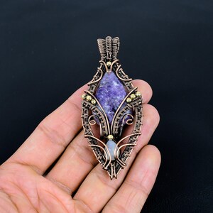 May include: A handcrafted copper wire pendant with a purple stone center and small gold-colored accents. The pendant has a teardrop shape with intricate wire wrapping and a small, light-colored stone at the bottom.