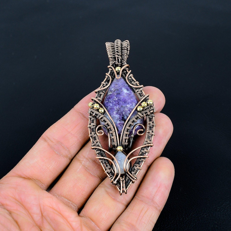 May include: A handcrafted copper wire pendant with a purple stone center and small gold-colored accents. The pendant has a teardrop shape with intricate wire wrapping and a small, light-colored stone at the bottom.