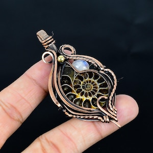 Puede incluir: Colgante artesanal con un amonita fosilizado envuelto en alambre de cobre. El colgante tiene un patr&oacute;n en espiral en tonos marrones y amarillos, con un peque&ntilde;o acento de piedra lunar y perla dorada. El trabajo del alambre es intrincado.