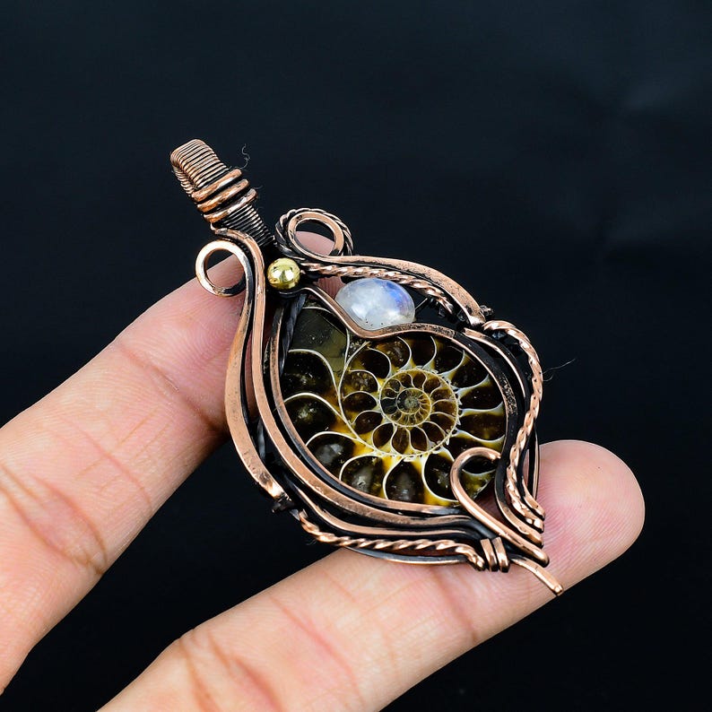 Puede incluir: Colgante artesanal con un amonita fosilizado envuelto en alambre de cobre. El colgante tiene un patr&oacute;n en espiral en tonos marrones y amarillos, con un peque&ntilde;o acento de piedra lunar y perla dorada. El trabajo del alambre es intrincado.