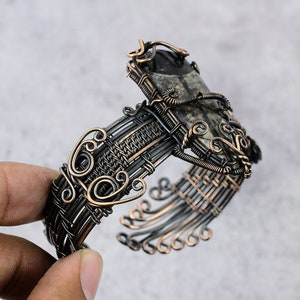 May include: A silver and copper wire wrapped bracelet with a dark gray and white stone set in the center. The bracelet is intricately woven with a decorative design.