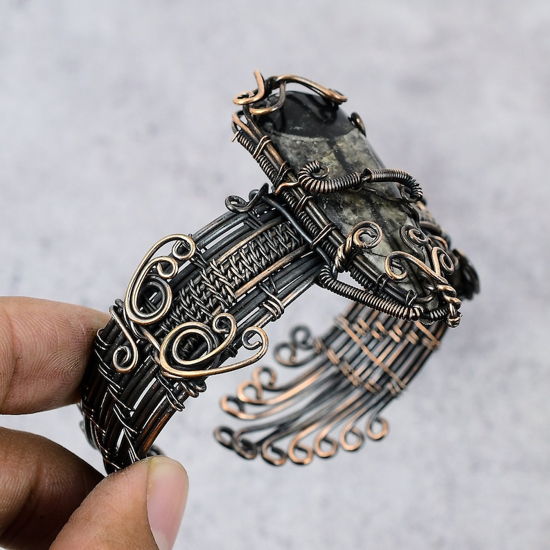 May include: A silver and copper wire wrapped bracelet with a dark gray and white stone set in the center. The bracelet is intricately woven with a decorative design.