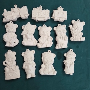 May include: A collection of unpainted ceramic ornaments in various shapes, including a train, train car, and teddy bears in different poses. Each ornament has a small hole for hanging.