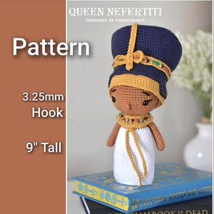 Puede incluir: Muñeca de ganchillo de la reina Nefertiti, de aproximadamente 23 cm de altura, con una cofia azul y dorada y un vestido blanco. La muñeca tiene una cara marrón y está adornada con un collar de cuentas. La imagen incluye el texto "Pattern" y "3.25mm Hook".