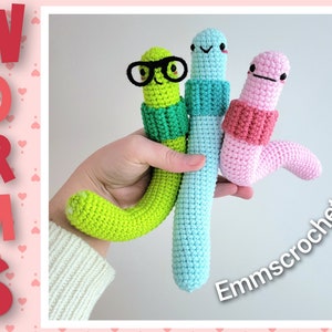 Puede incluir: Tres peluches de gusanos de crochet en verde, azul y rosa. Cada gusano tiene un gorro de crochet y una cara. El gusano verde lleva gafas. El texto "Worms" está en letras rojas en el lado izquierdo de la imagen. El texto "Emmcrochet" está en letras negras en el lado derecho de la imagen.