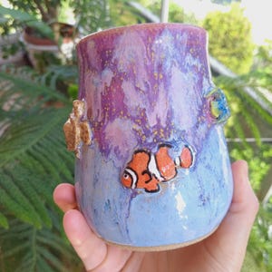 Può includere: Tazza in ceramica fatta a mano con un design a tema oceanico. La tazza presenta un pesce pagliaccio, una tartaruga e una colorata smaltatura a vortice nei toni del blu, viola e oro. La tazza è tenuta in mano.
