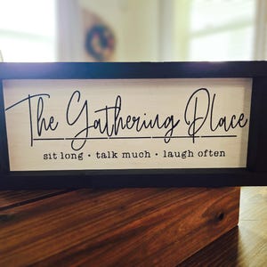 May include: A rectangular wooden sign with a black frame. The sign has a light-colored background with the words "The Gathering Place" in a cursive font. Below the main text are the words "sit long • talk much • laugh often" in a smaller, sans-serif font.