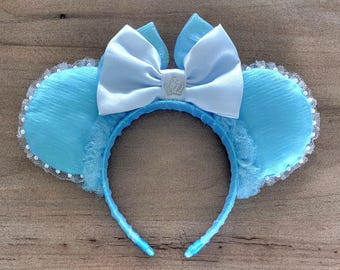 May include: A light blue headband with mouse ears. The ears are a textured light blue fabric with a white lace trim and small white pearls. A large white satin bow with a small silver crown in the centre sits atop the headband.