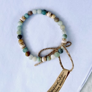 May include: A green, brown and white beaded bracelet with a gold accent. The bracelet is tied with a brown string and has a tag that reads "Breath and Collective Aroma Bracelet".