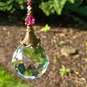 May include: A faceted crystal ball suncatcher with a brass-colored filigree base and a pink crystal accent. The suncatcher is suspended by a string of colorful beads, reflecting light and creating a rainbow effect. The background is blurred green foliage.