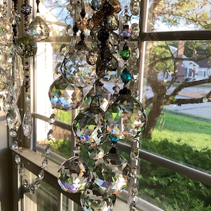May include: A crystal suncatcher with multiple faceted crystal balls hanging from a chain. The suncatcher is hanging in a window with a view of green trees and grass.