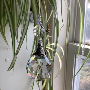 May include: A faceted crystal ball suncatcher with a silver-colored metal cap and hanging beads. The crystal reflects light, creating a rainbow effect. The suncatcher is suspended in front of green and white striped leaves.