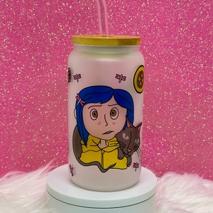 May include: A white frosted glass tumbler with a bamboo lid and a clear drinking straw. The tumbler features a colorful illustration of a girl with blue hair and a black cat. The girl is wearing a yellow jacket and has a worried expression. The tumbler has a pink background with pink dragonfly illustrations.