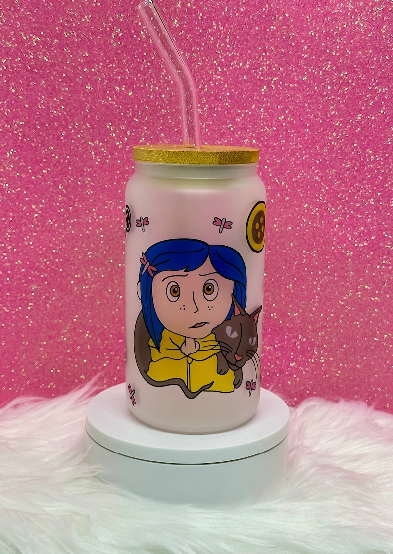 May include: A white frosted glass tumbler with a bamboo lid and a clear drinking straw. The tumbler features a colorful illustration of a girl with blue hair and a black cat. The girl is wearing a yellow jacket and has a worried expression. The tumbler has a pink background with pink dragonfly illustrations.