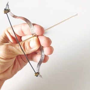 May include: A miniature silver bow and arrow set. The bow is made of a thin, curved metal strip with a black string. The arrow is a wooden stick with a white tip. The set is held in a hand against a white background.