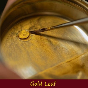 May include: A small metal container with a gold liquid inside. A brush is being used to stir the liquid. The container is labeled "Gold Leaf".