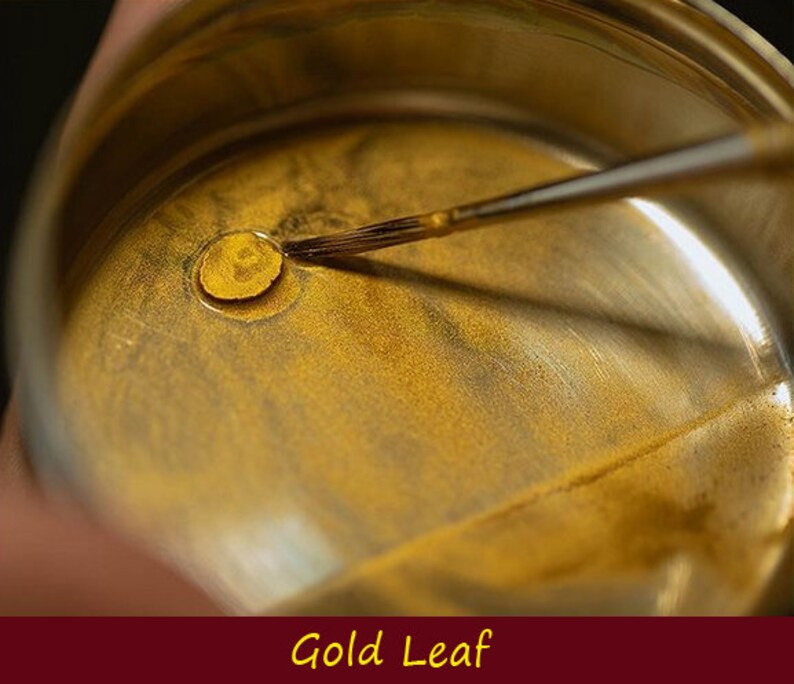 May include: A small metal container with a gold liquid inside. A brush is being used to stir the liquid. The container is labeled "Gold Leaf".