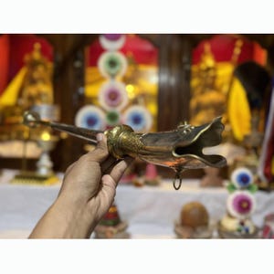 May include: A long, metallic ritual horn featuring an ornate, flared mouthpiece shaped like an open creature's mouth. The body of the horn is slender and tapers toward a narrow end, with intricate engraved patterns decorating the brass-toned surface.