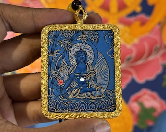 May include: A rectangular pendant featuring a blue and gold illustration of a seated figure in a meditative pose, surrounded by bamboo and floral motifs. The image is encased in an ornate, textured gold-coloured metal frame with a top bail.