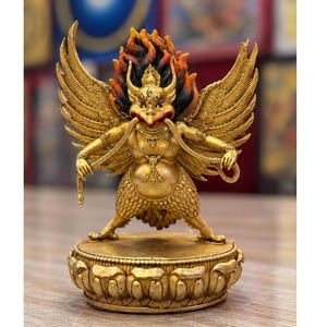 May include: A golden statue of a winged deity with a bird-like face, horns, and a third eye, standing on a lotus base. The figure has large, detailed wings, holds a snake in its hands, and is surrounded by a flame-like aura in red, orange, and black.