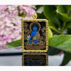 May include: A rectangular gold-toned pendant featuring a central illustration of a blue Buddha figure seated on a lotus throne. The figure is surrounded by gold-colored clouds against a black background, all encased in an ornate, textured gold frame.