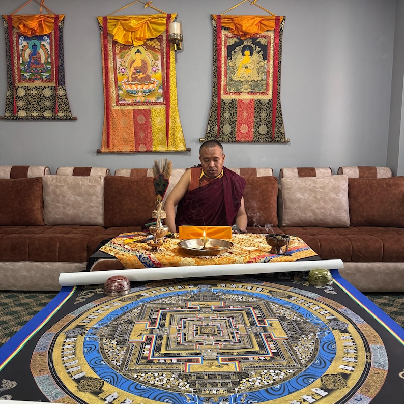 May include: A monk in a red and maroon robe sits in front of a table with a golden mandala on a blue background. The mandala is surrounded by a black border. The monk is holding a small golden object in his right hand. There are three golden statues of Buddha on the wall behind the monk.