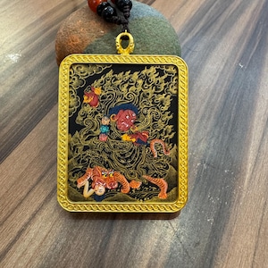 May include: A gold-colored rectangular pendant with an intricate design featuring a red-faced figure, a tiger, and other figures on a black background. The pendant is suspended from a black cord with red and black beads.