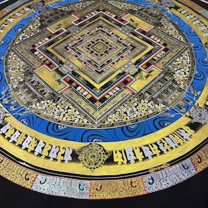 May include: A circular mandala with a square center, surrounded by a blue and gold border. The mandala is painted in gold, black, red, and blue. The center square is filled with intricate patterns and designs. The border is decorated with a repeating pattern of flowers and leaves.