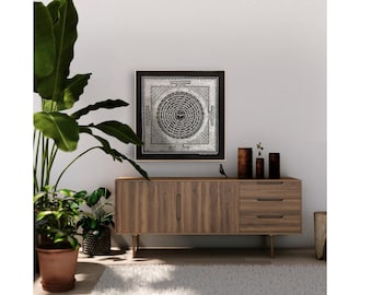 May include: A black framed wall art print with a silver mandala design and intricate details. The print is hanging on a white wall above a wooden cabinet with three drawers.