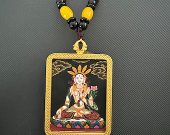 May include: A rectangular pendant necklace featuring a seated figure with a gold frame. The figure is depicted with a serene expression, wearing a crown and holding a lotus flower. The necklace has a brown cord with yellow and black beads.