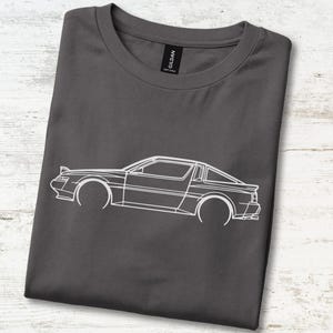 May include: A charcoal grey crew neck t-shirt featuring a white line art illustration of a sports car in profile. The Gildan brand label is visible at the inner neckline. The design depicts the car's silhouette with pop-up headlights.