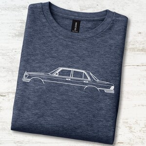 May include: A dark heather navy blue crew neck t-shirt featuring a white line art graphic of a classic sedan car on the front. The shirt has a black Gildan brand label at the inner neckline and is made of a soft, heathered fabric material.
