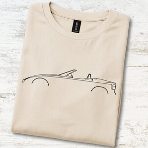 May include: A beige short-sleeved t-shirt featuring a minimalist black line art illustration of a convertible car in profile. The shirt has a crew neckline and a black Gildan brand label visible at the inner collar, centred on the fabric.