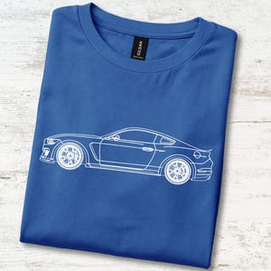 Può includere: Una t-shirt a maniche corte color blu reale con una grafica a linee bianche che raffigura un'auto sportiva di profilo. Il capo presenta un girocollo e un'etichetta nera del marchio Gildan visibile sul colletto interno.