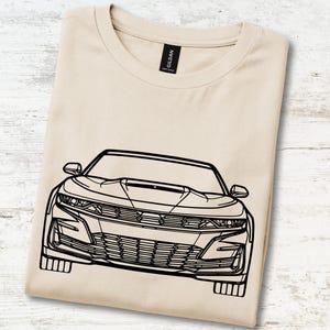 Pode incluir: Uma t-shirt bege de gola redonda com uma ilustração de linhas pretas da vista frontal de um carro desportivo. O gráfico retrata os faróis, a grelha e o capô do veículo num estilo de contorno minimalista. Etiqueta preta na gola.
