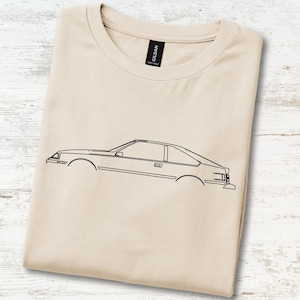 May include: A sand-coloured t-shirt featuring a black line art illustration of a car in profile. The shirt has a crew neckline and a black Gildan brand label at the inner collar. The design depicts the side silhouette of a vehicle without wheels.