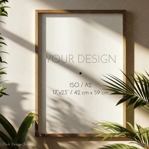 May include: A wooden frame mockup with a white mat and a white background. The frame is sitting on a beige wall with a green plant in the foreground. The text "YOUR DESIGN" is printed on the mat, along with the dimensions "17"x23" / 42 cm x 59 cm" and "ISO/A2".