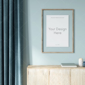May include: A light blue wall with a wooden frame poster mockup, a light blue curtain, and a wooden cabinet with a carved design. The poster mockup has the text "Your Design Here" in the center.