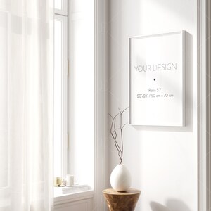May include: A white frame with the text "YOUR DESIGN" and the dimensions "20'x28' / 50 cm x 70 cm" in black text. The frame is hanging on a white wall with a white vase and a wooden stool in the foreground.