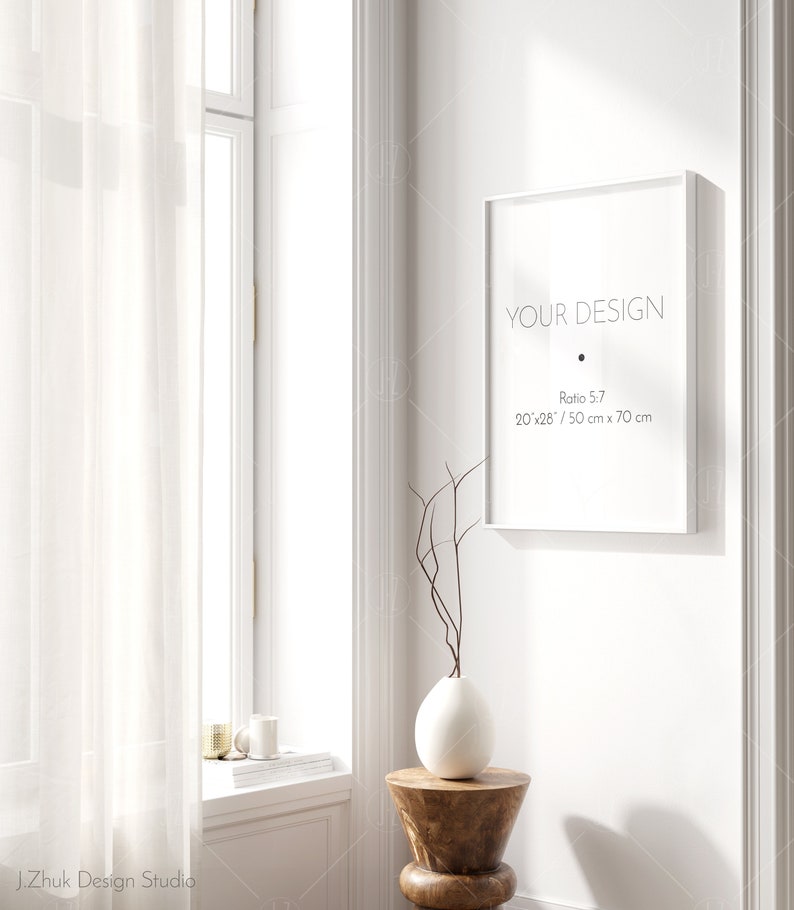 May include: A white frame with the text "YOUR DESIGN" and the dimensions "20'x28' / 50 cm x 70 cm" in black text. The frame is hanging on a white wall with a white vase and a wooden stool in the foreground.