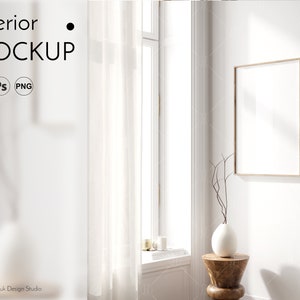 May include: A white vase with a dried branch sits on a wooden stool in front of a large, framed picture hanging on a white wall. The picture is blank and the frame is light brown. A white sheer curtain hangs in front of a window with sunlight streaming through it.
