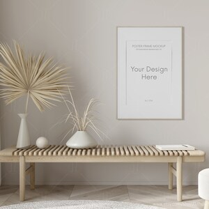 Puede incluir: Un banco de madera beige con una maqueta de póster enmarcado en blanco encima. El póster tiene el texto "Your Design Here". Hay dos plantas secas en jarrones blancos sobre el banco. El banco está frente a una pared de color beige claro.