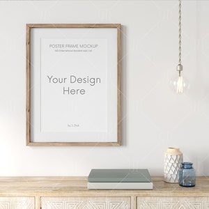 May include: A wooden frame mockup with the text "POSTER FRAME MOCKUP 150 International standard size / A2" and "Your Design Here" by 1 Zhuk. The frame is hanging on a white wall above a wooden cabinet with a light blue book and two decorative vases.