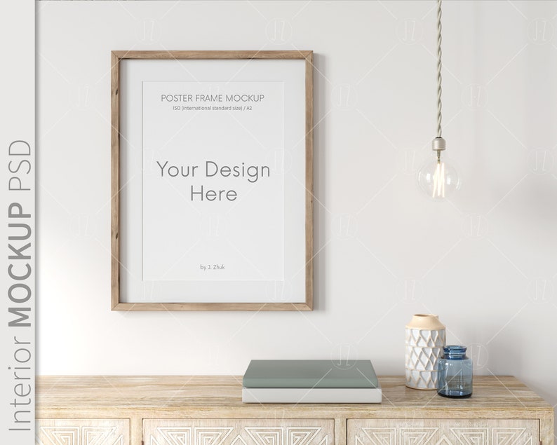 May include: A wooden frame mockup with the text "POSTER FRAME MOCKUP 150 International standard size / A2" and "Your Design Here" by 1 Zhuk. The frame is hanging on a white wall above a wooden cabinet with a light blue book and two decorative vases.