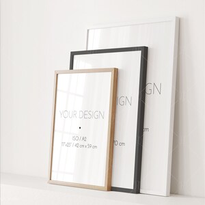 May include: Three picture frames of different sizes and colors, with the smallest frame in the front and the largest frame in the back. The smallest frame is made of natural wood and has the text "YOUR DESIGN" and "ISO/A2 17x25/ 42 cm x 59 cm" printed on it. The middle frame is black and the largest frame is white.
