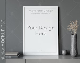 May include: A white poster frame mockup with the text "POSTER FRAME MOCKUP ISO (international standard size) / A2 Your Design Here by J. Zhuk" on a gray wall. The frame is sitting on a gray table with a stack of books and a gray vase with dried flowers.
