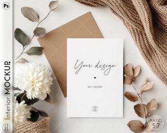 May include: A white card mockup with the text "Your design" and "5x7 inches" on a white surface with a brown envelope, a white flower, and a brown knitted scarf.