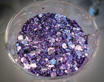 May include: A glass goblet filled with iridescent purple and silver glitter. The glitter is a mix of small and large pieces, creating a sparkling effect.