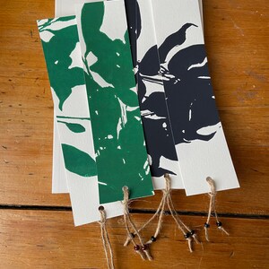 Pode incluir: Quatro marcadores de página com desenhos abstratos pretos e verdes impressos em cartão branco. Cada marcador de página tem um furo perfurado na parte superior e é amarrado com um cordão castanho com contas castanhas escuras e pretas.